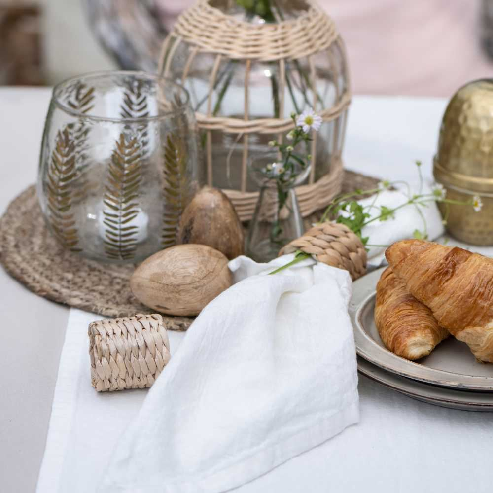 Frühstückstisch mit weißer Leinentischdecke, Croissants auf Steingut, Glaswindlicht und kleinem Rattankorb