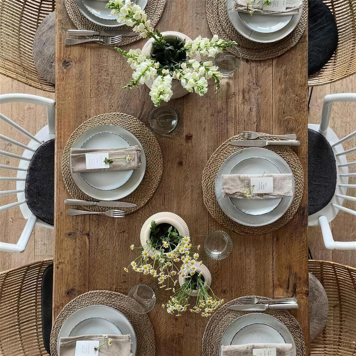 Gedeckter Holztisch von oben mit Steingut-Tellern auf Jute-Tischsets, Leinenservietten und kleinen Vasen mit Kamille
